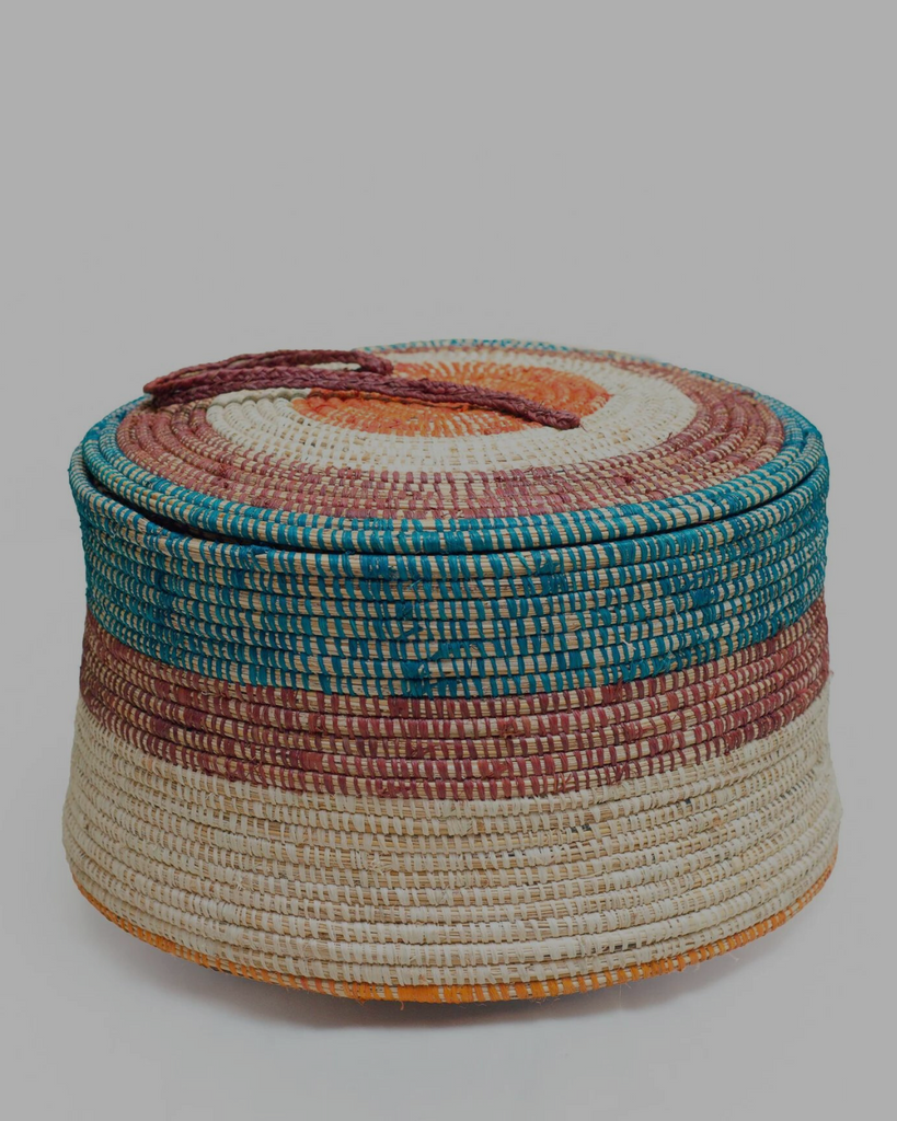 woven flat-lidded basket with off-white, brown, and light blue banding, with an orange circle at the top and leather strap handle